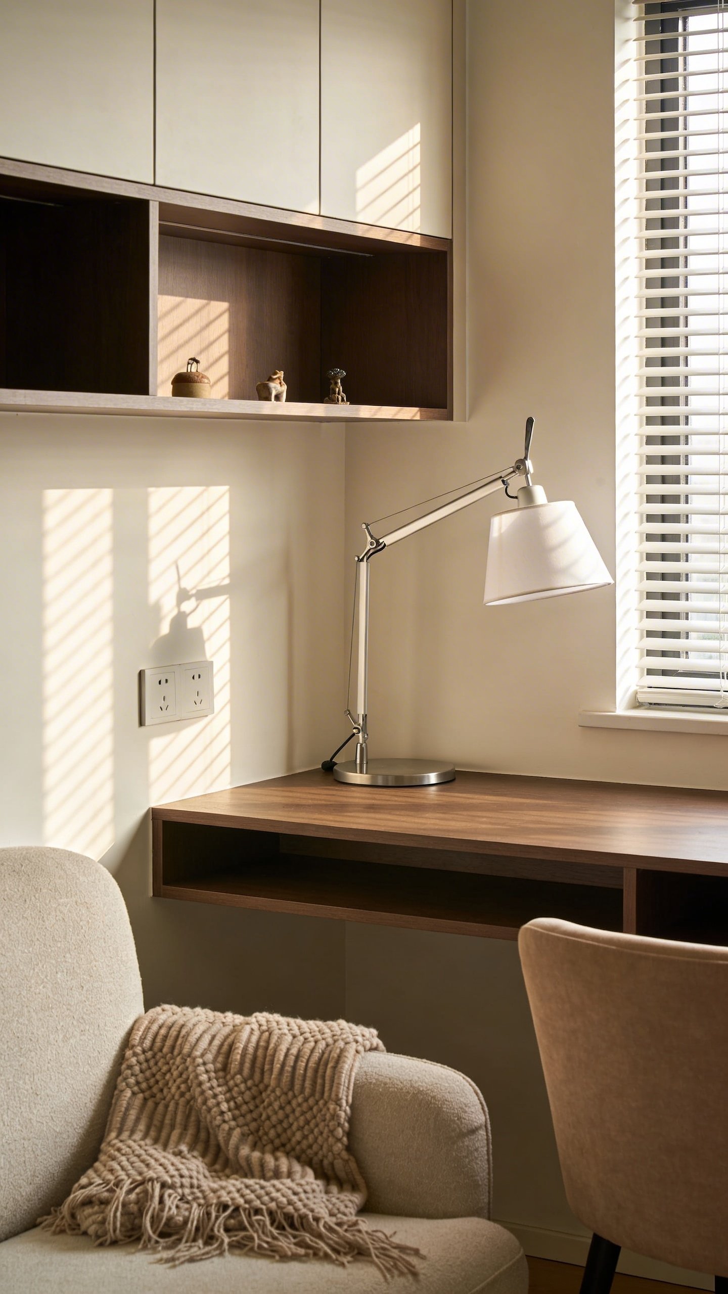 A small home office niche created within a living room: a compact desk under a window, a wall-mounted shelf unit, a task lamp, and a cozy chair with a throw, all arranged to form a dedicated but unobtrusive desk zone.