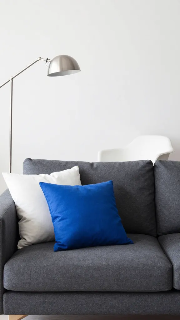 A playful yet sophisticated scene: a dark gray sofa dressed with charcoal, graphite, and white pillows, plus a cobalt blue accent pillow as the sole color pop; a mid-century chair in white, a sleek metal floor lamp, and a clean, bright backdrop.