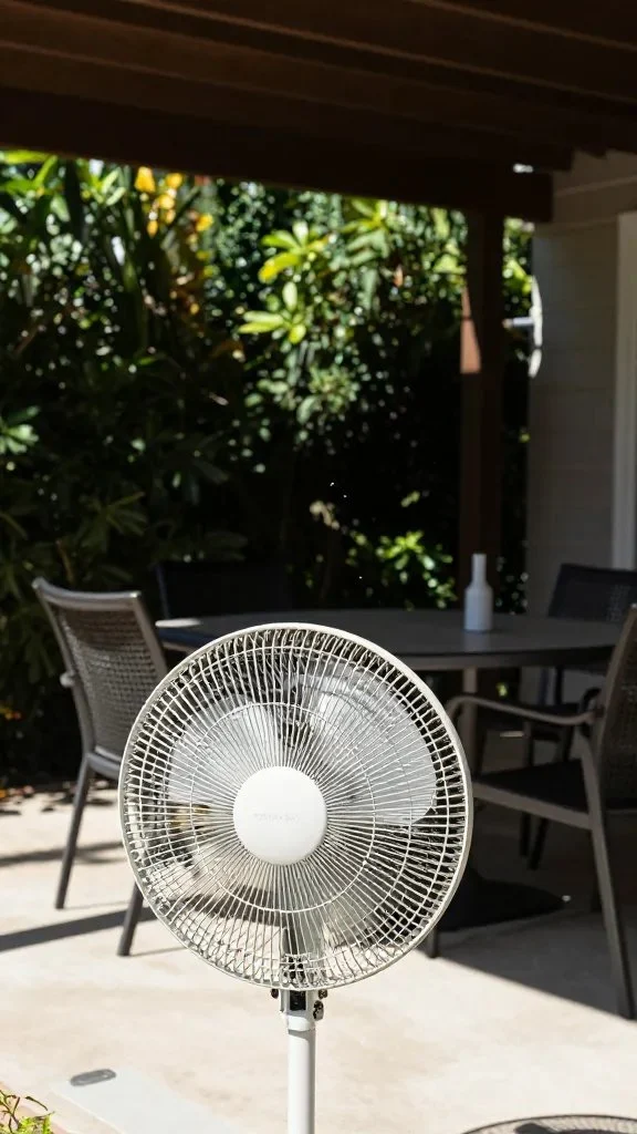 A sunlit patio with a fan in use, outdoor furniture pushed aside, and a few mosquitoes visible in the air near the edge of the shade line under a pergola.