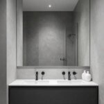 Create a hyper-realistic, high-resolution bathroom interior featuring a Monochrome Minimal with Mirror Wall Drama concept: a compact modern bathroom with a full-length floor-to-ceiling mirror spanning one entire wall, reflecting a crisp grayscale palette of charcoal, dove gray, and white. Include a recessed vanity with handle-free drawers in charcoal finish, subtle black metal hardware for a pop, and a minimalist glass shower enclosure with clear glass. The countertops should be clear and uncluttered, with a few white porcelain vessels kept to a minimum. Emphasize clean lines, soft diffused lighting, and a seamless, mirror-dominated wall that visually expands the space. The main subject from the article title—modern minimalist bathroom design—should be represented through the chic, restrained aesthetic, precision textures (matte concrete, smooth porcelain, glass), and a polished yet understated atmosphere. No text or branding in the scene.