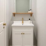 A small bathroom with a cohesive color palette of warm whites, soft gray walls, and sage accents; a muted accent wall, white fixtures, warm wood shelving, and pebble-textured bath mat, with brass fixtures and no clutter.